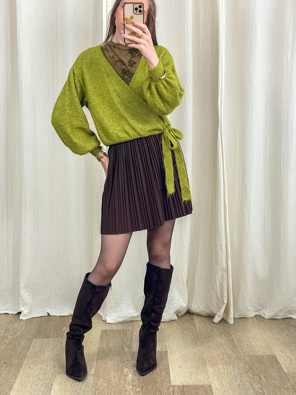 Jupe courte plissée marron taille haute femme portée avec pull cache-cœur vert et bottes hautes