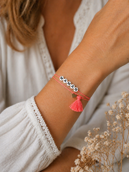 Bracelet affectif Maman porté au poignet — cordon corail, perles lettres blanches et pompon corail avec breloque cœur doré