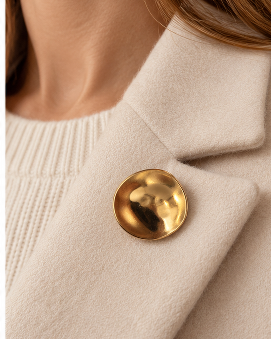 Broche ronde dorée effet martelé brillant portée sur col de manteau beige, accessoire bijou femme élégant