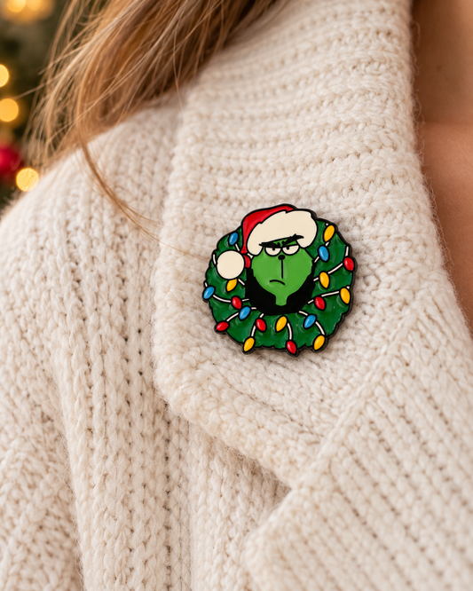 Pin's Grinch Noël en émail vert avec couronne lumineuse colorée et bonnet rouge, accessoire fantaisie fêtes porté sur pull